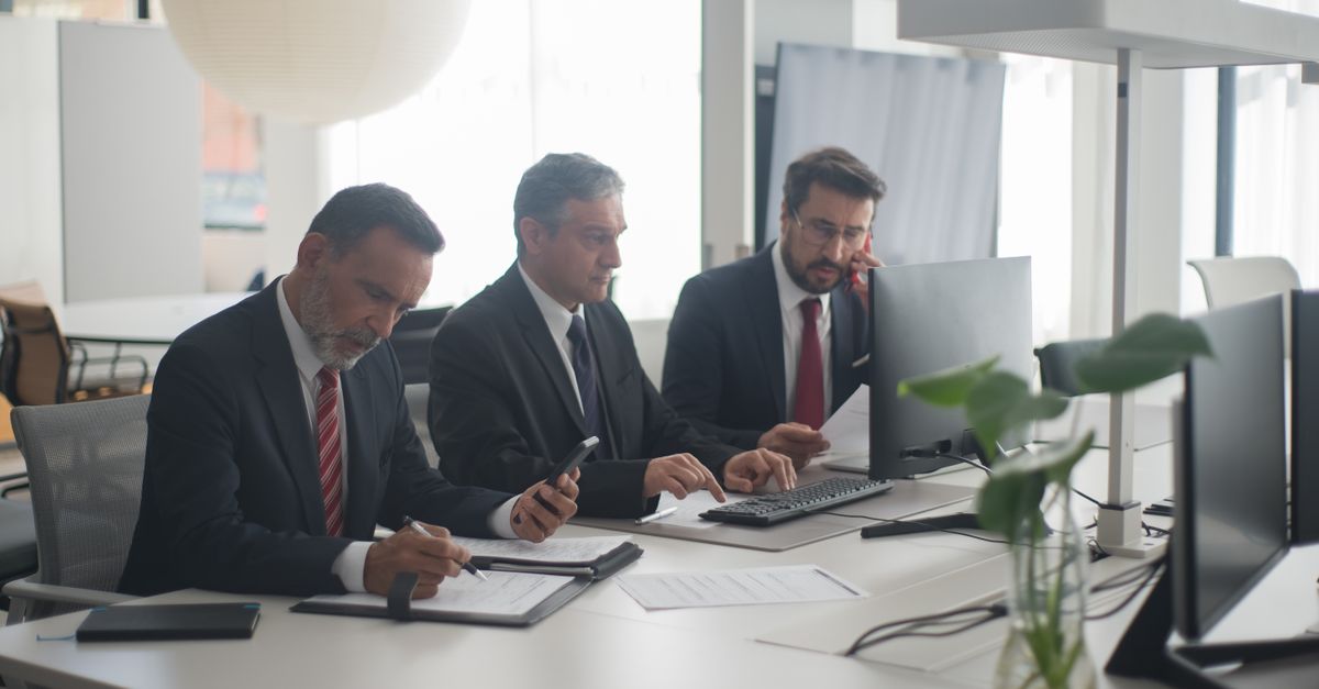 Compare Debt Solutions Three men in suits collaborating at a modern office desk with computers and documents.